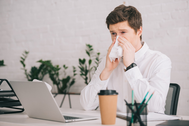 schöner Geschäftsmann niest in Taschentuch, während er neben Laptop im Büro sitzt  - Foto, Bild