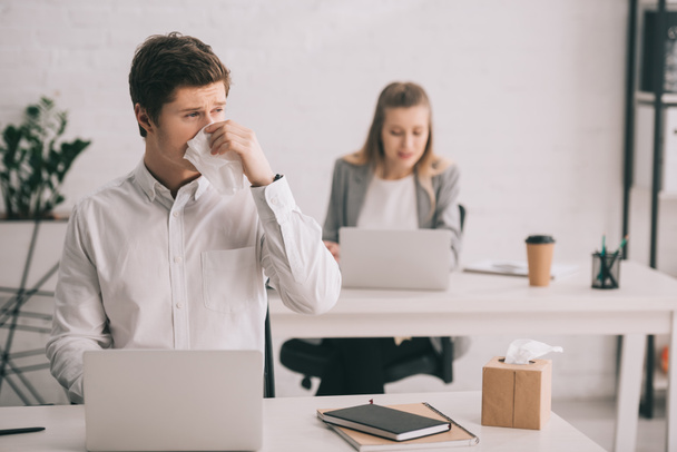 selective focus of businessman sneezing in tissue while using laptop near businesswoman in office  - Foto, Imagem