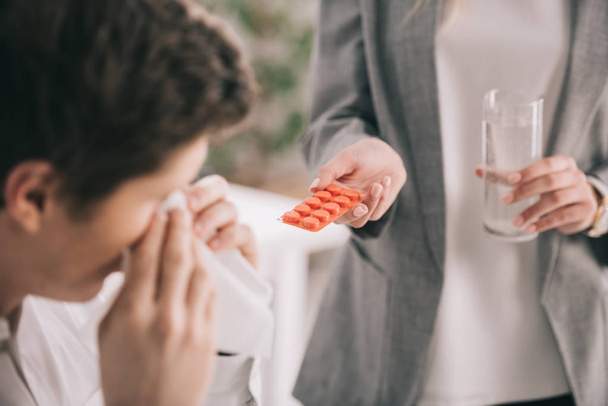 Ausgeschnittene Ansicht einer Frau, die Tabletten und ein Glas Wasser in der Nähe eines Mitarbeiters hält, der in Gewebe niest  - Foto, Bild