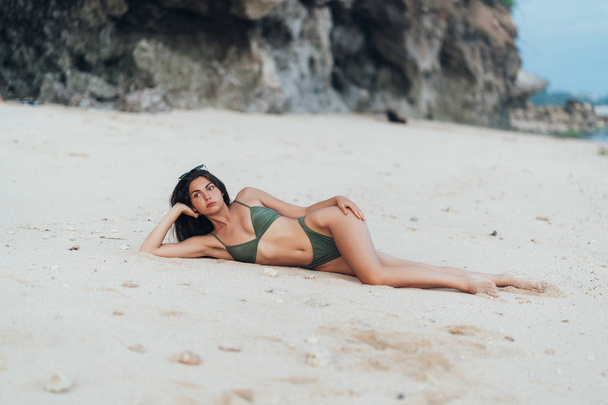 Beautiful tanned girl in green swimwear and sunglasses posing on beach with rock on background - 写真・画像