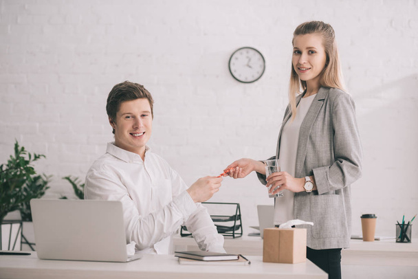 cheerful businesswoman holding pills and glass of water near happy colleague in office  - Foto, imagen