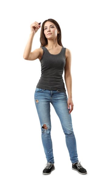 Young woman in gray sleeveless top and blue jeans standing and posing with marker in hand as if writing on invisible board isolated on white background. - Zdjęcie, obraz