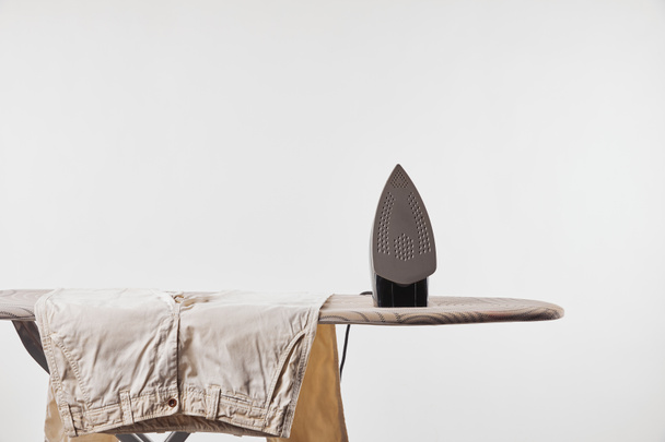 Ironing board, beige pants and iron isolated on grey - Photo, Image