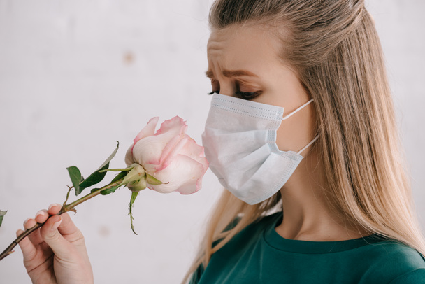 ξανθιά γυναίκα με αλλεργία στη γύρη φορώντας ιατρική μάσκα και μυρίζει τριαντάφυλλο  - Φωτογραφία, εικόνα