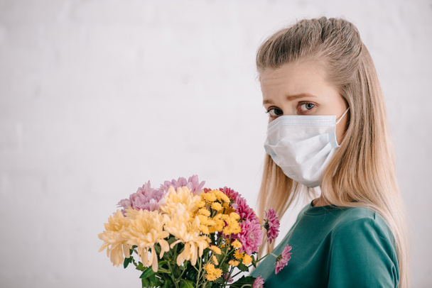 blonde woman with pollen allergy wearing medical mask and holding flowers while looking at camera  - Fotó, kép