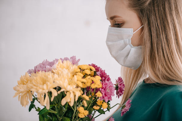 Blonde Frau mit Pollenallergie trägt medizinische Maske und betrachtet Blumenstrauß  - Foto, Bild