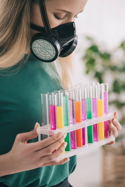 blond meisje in respiratoire masker kijken naar kleurrijke reageerbuisjes  - Foto, afbeelding