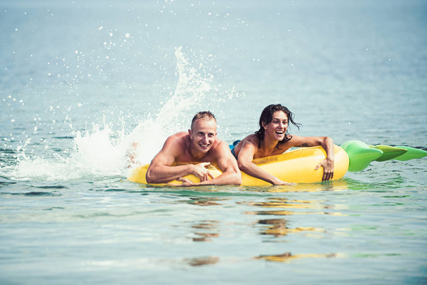 Happy young couple swimming and laughing on air mattress. Couple vacation concept. Man and woman on honeymoon, swim on pineapple shaped mattress in sea. Summer accessories for rest yellow mattress - Photo, Image