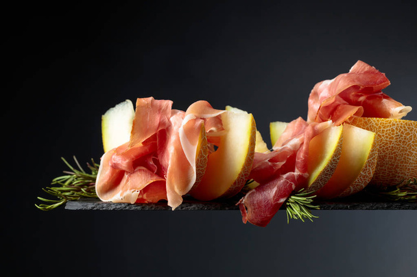 Prosciutto with melon and rosemary  on a black background. - Photo, Image