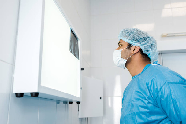 vista lateral del médico en gorra médica y guante de látex mirando rayos X en la clínica
  - Foto, Imagen