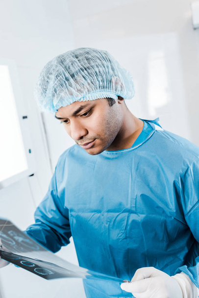 όμορφος γιατρός σε ιατρικό καπάκι και ομοιόμορφη κρατώντας ακτινογραφία στην κλινική  - Φωτογραφία, εικόνα