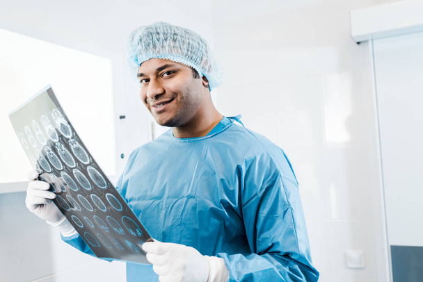 smiling doctor in uniform and medical cap holding x-ray in and looking at camera  - Foto, Imagem