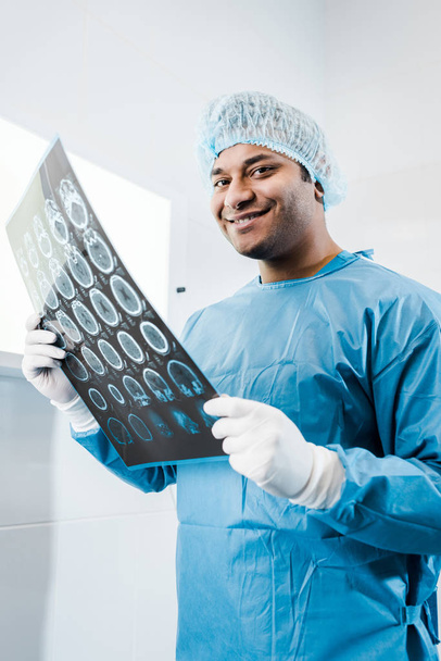 smiling doctor in uniform and medical cap holding x-ray in and looking at camera  - Foto, Imagen
