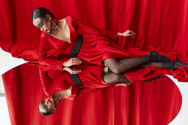 elegante und sexy schöne junge Frau in rotem Seidenkleid und schwarzer Spitze im Gesicht, die in den großen runden Spiegel auf dem Boden blickt. roter Vorhang auf Hintergrund - Foto, Bild