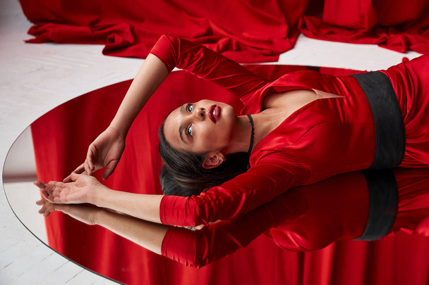 Elegant and sexy beautiful young brunette woman in red silky dress laying on big round mirror. Red curtain on background - Foto, immagini