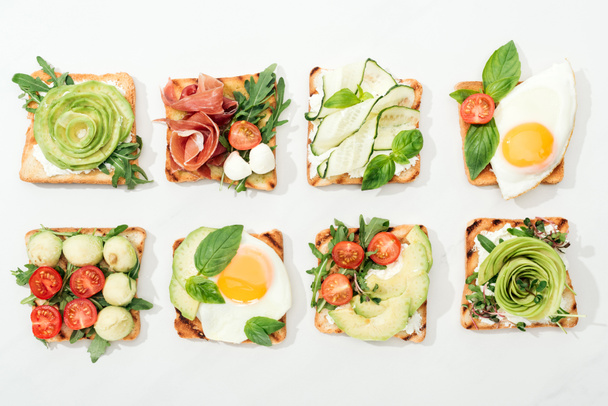 Top view of toasts with cut vegetables and prosciutto on white surface - 写真・画像