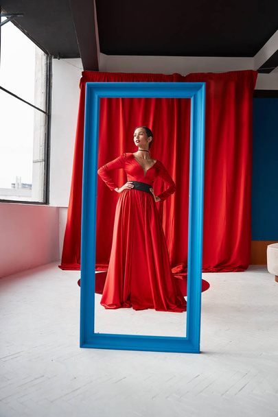 Young sexy woman in long red dress standing behind blue frame. Red curtains on background.  - Foto, afbeelding