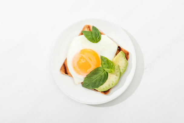 Top view of toast with fried egg and cut avocado on plate on white surface - Photo, Image
