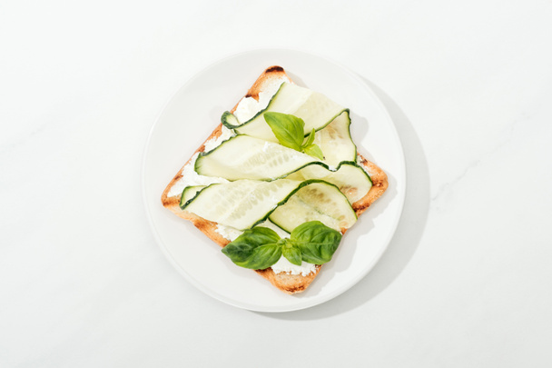 Top view of toast with sliced cucumber and basil on plate on white surface - Foto, Imagem