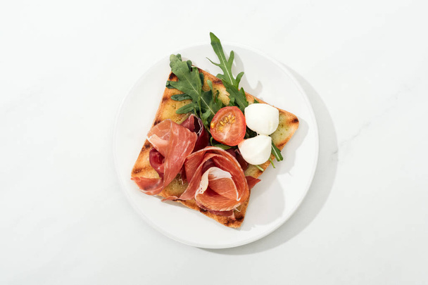 Top view of toast with cherry tomato, arugula and prosciutto on plate on white surface - Valokuva, kuva