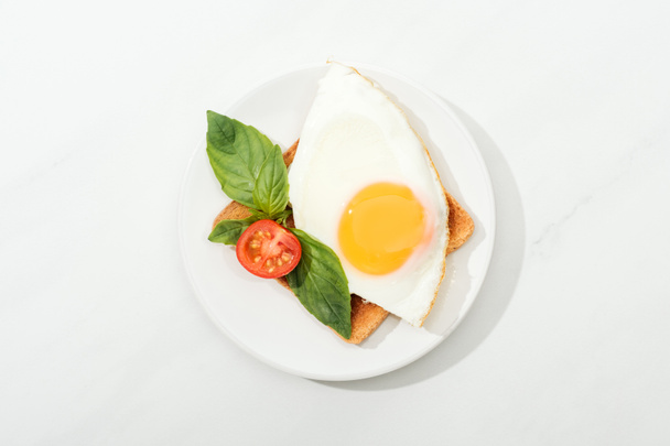 Toast met gebakken ei, basilicum en gesneden tomaat cherry op plaat op witte oppervlak (bovenaanzicht) - Foto, afbeelding