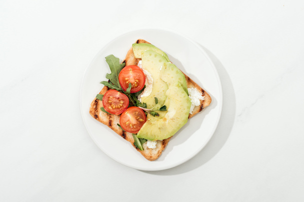 Draufsicht auf Toast mit Kirschtomaten und Avocado auf Teller auf weißer Oberfläche - Foto, Bild