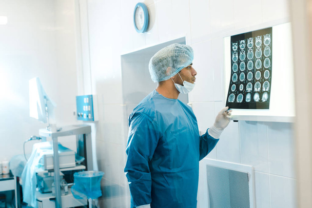 vista lateral del médico guapo en uniforme y máscara médica celebración de rayos X en la clínica
  - Foto, Imagen
