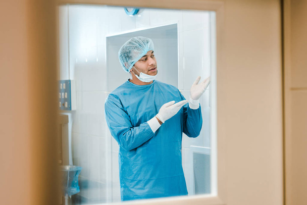 guapo doctor en uniforme y máscara médica poniéndose guante de látex y mirando hacia otro lado
  - Foto, Imagen