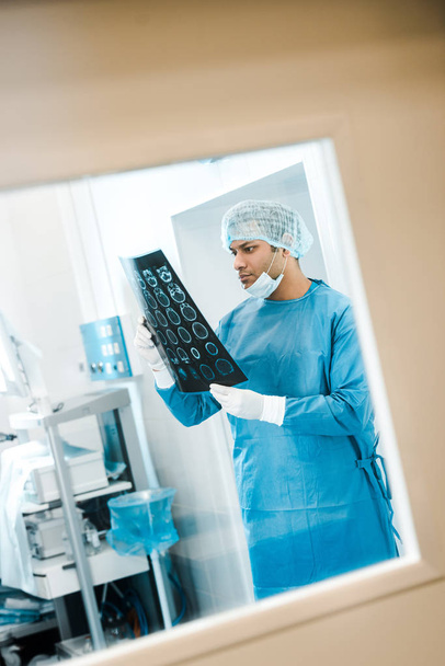 όμορφος γιατρός με στολή και ιατρικό καπάκι κοιτάζοντας ακτίνες χ στην κλινική  - Φωτογραφία, εικόνα