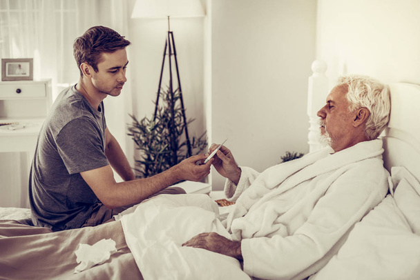 Young-adult son handing the thermometer to his aged ill father - Fotografie, Obrázek