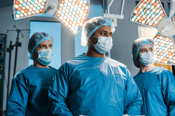 médicos y enfermeras en uniformes y máscaras médicas mirando hacia otro lado en el quirófano
 - Foto, Imagen