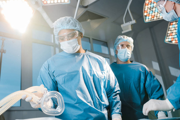 лікарі і медсестра в уніформі і медичні шапки тримають маску в операційній кімнаті
 - Фото, зображення