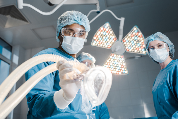 médico e enfermeiro em uniformes e bonés médicos segurando máscara na sala de cirurgia
 - Foto, Imagem