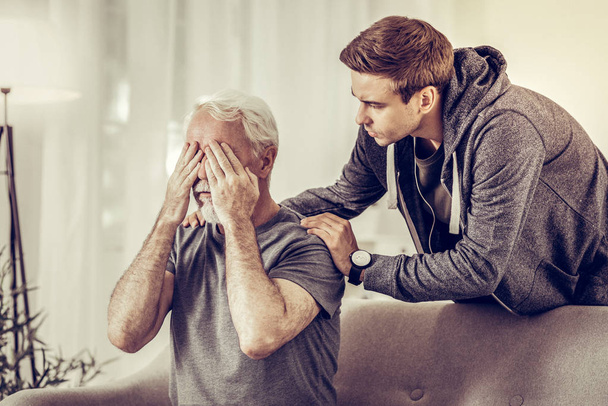 Young appealing upset man calmingly hugging ill sorrowful grey-haired grandfather - Foto, Imagen