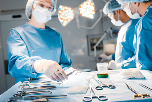 selective focus of doctors doing operation and nurse in uniform and medical cap holding scalpel  - Photo, Image