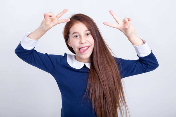 Portrait of a cheerful girl who shows tongue and fingers sign V on a light background. Cheerful schoolgirl indulge and weigh. - Foto, immagini