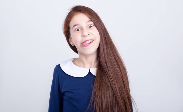 Portrait of a girl with a funny facial expression on a light background. Cheerful schoolgirl makes faces and shows her tongue. - Foto, imagen
