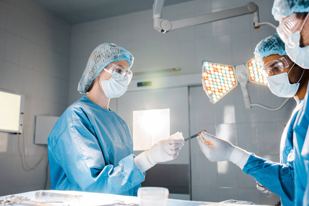 nurse in uniform and medical cap giving scalpel to doctor in operating room  - Photo, Image