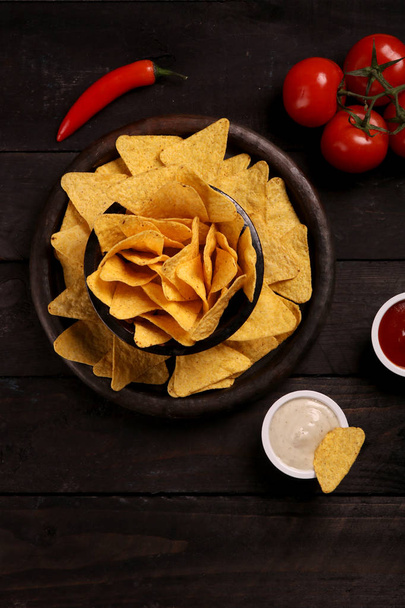 Мексиканские начос в тортилье из деревянного кубка с чипсами с соусом чили, соусом сальсы и сыром, видом сверху и черным фоном
 - Фото, изображение