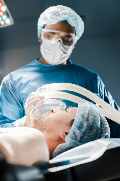 επιλεκτική εστίαση του γιατρού σε ομοιόμορφη και ιατρικό καπάκι βάζοντας μάσκα στον ασθενή  - Φωτογραφία, εικόνα