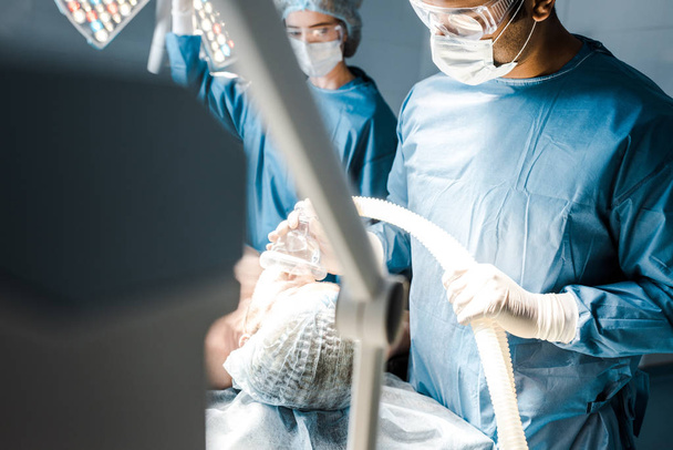 selective focus of surgeon in uniform and goggles putting mask on patient  - Photo, Image