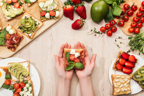 vue du dessus de la femme tenant un toast aux tomates cerises et au prosciutto sur une table en bois avec de la verdure et des ingrédients
 - Photo, image
