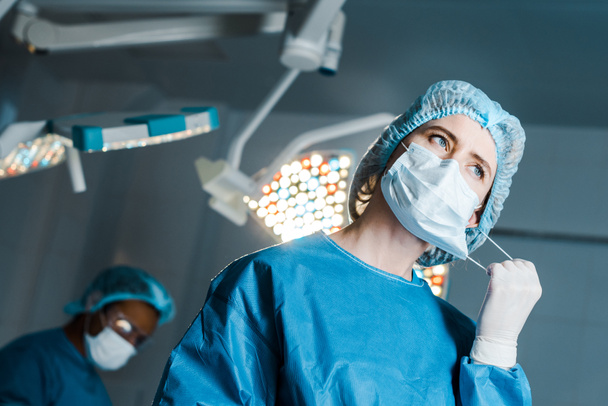 selective focus of nurse in uniform putting off medical mask in operating room - Photo, Image
