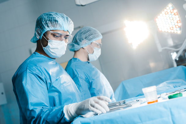 ošetřovatelka a chirurg v uniformě a lékařské masce, držící lékařské vybavení  - Fotografie, Obrázek