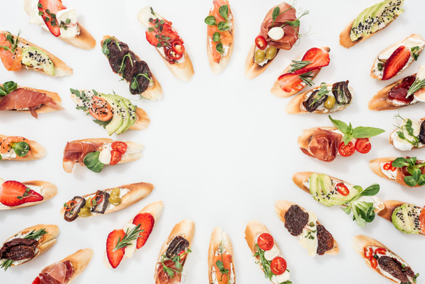 vista superior de marco redondo hecho de bruschetta italiana con salmón, jamón, hierbas y varias frutas con verduras en blanco
 - Foto, Imagen
