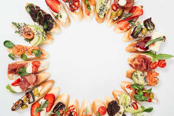 top view of round frame made of delicious italian bruschetta with salmon, prosciutto, herbs and various fruits with vegetables - Fotó, kép