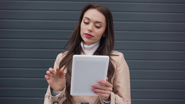 smiling elegant young woman in beige trench coat using digital tablet in windy weather - Materiał filmowy, wideo