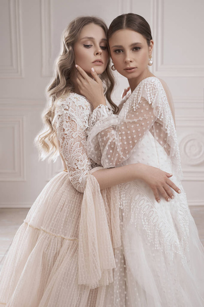 gorgeous blonde and brunette women in fashionable long evening dress hugging while posing in white studio interior  - Zdjęcie, obraz
