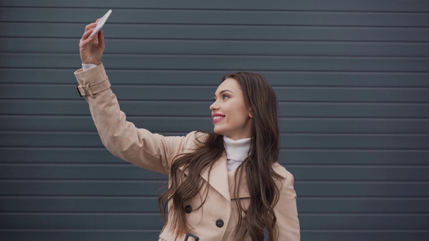 sonriente mujer elegante en gabardina beige tomando selfie en el teléfono inteligente en tiempo ventoso
 - Imágenes, Vídeo