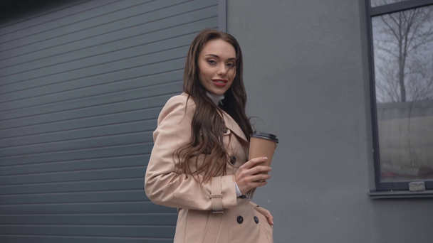 smiling elegant woman in beige trench coat walking with coffee to go near building - Materiał filmowy, wideo